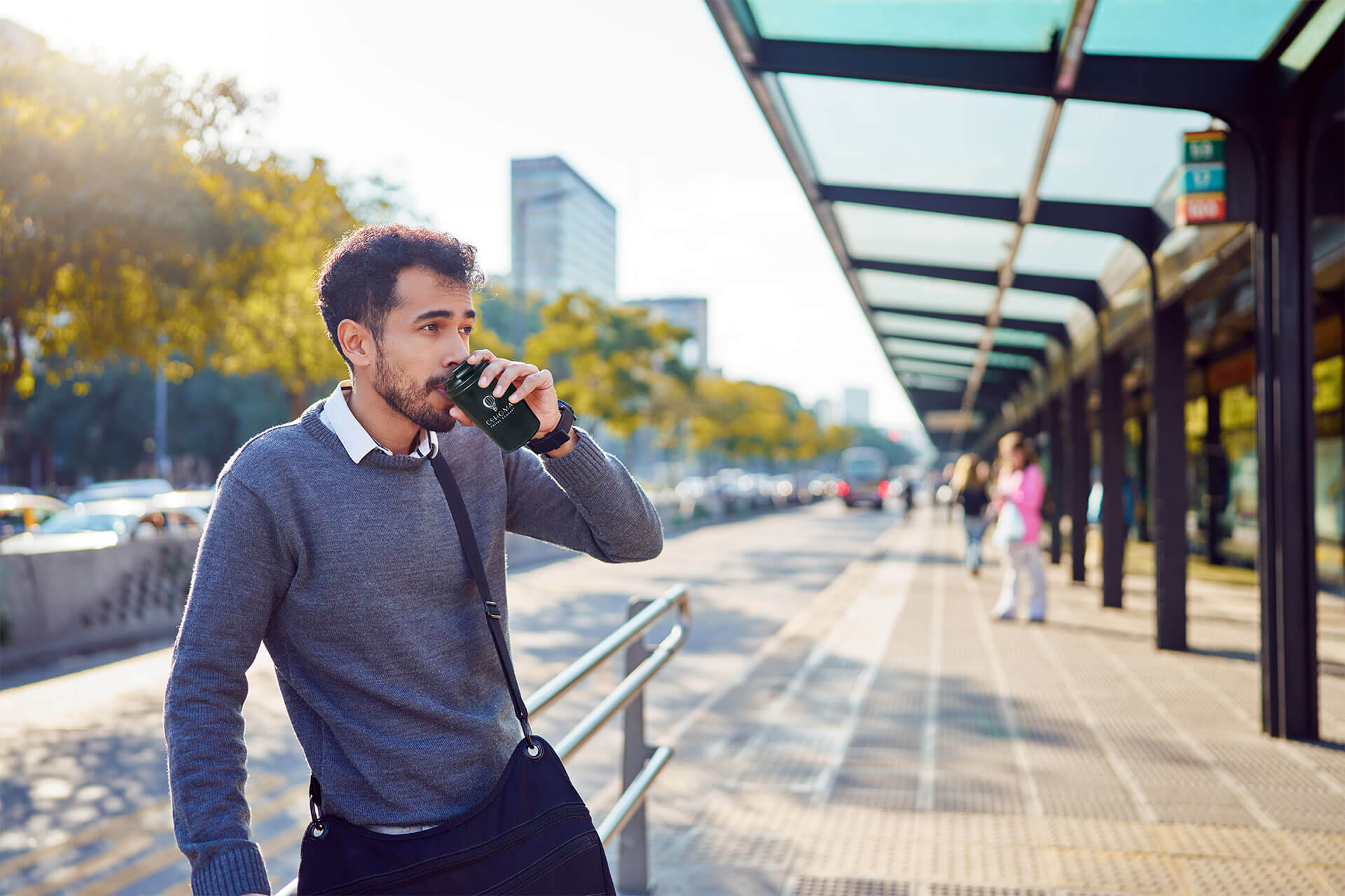 Man sipping from a Cohoma Coffee hot tumblr while standing at a sunny bus stop, wearing a gray sweater and crossbody bag