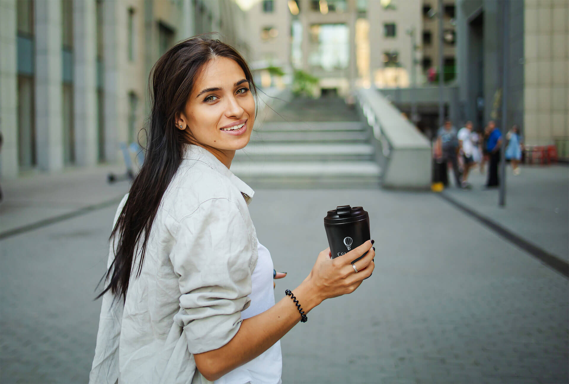 Smiling woman holding a Cohoma Coffee hot tumblr, walking in an urban setting with modern buildings in the background