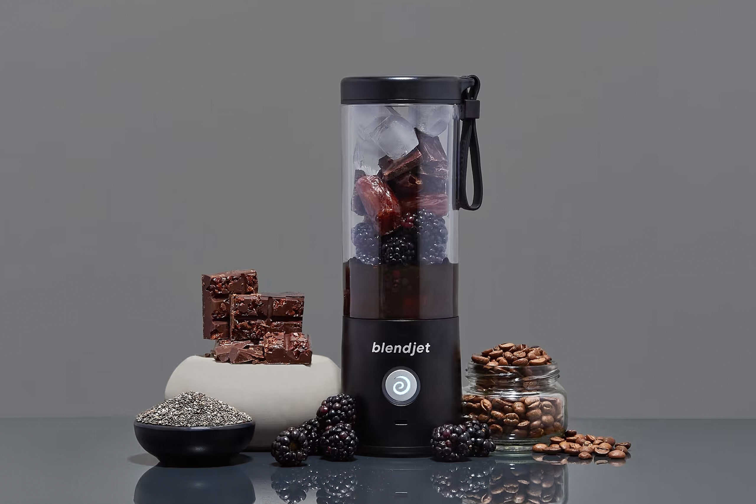 BlendJet 2 portable blender filled with blackberries, chocolate, and ice, surrounded by chia seeds, coffee beans, and dark chocolate pieces on a sleek gray surface