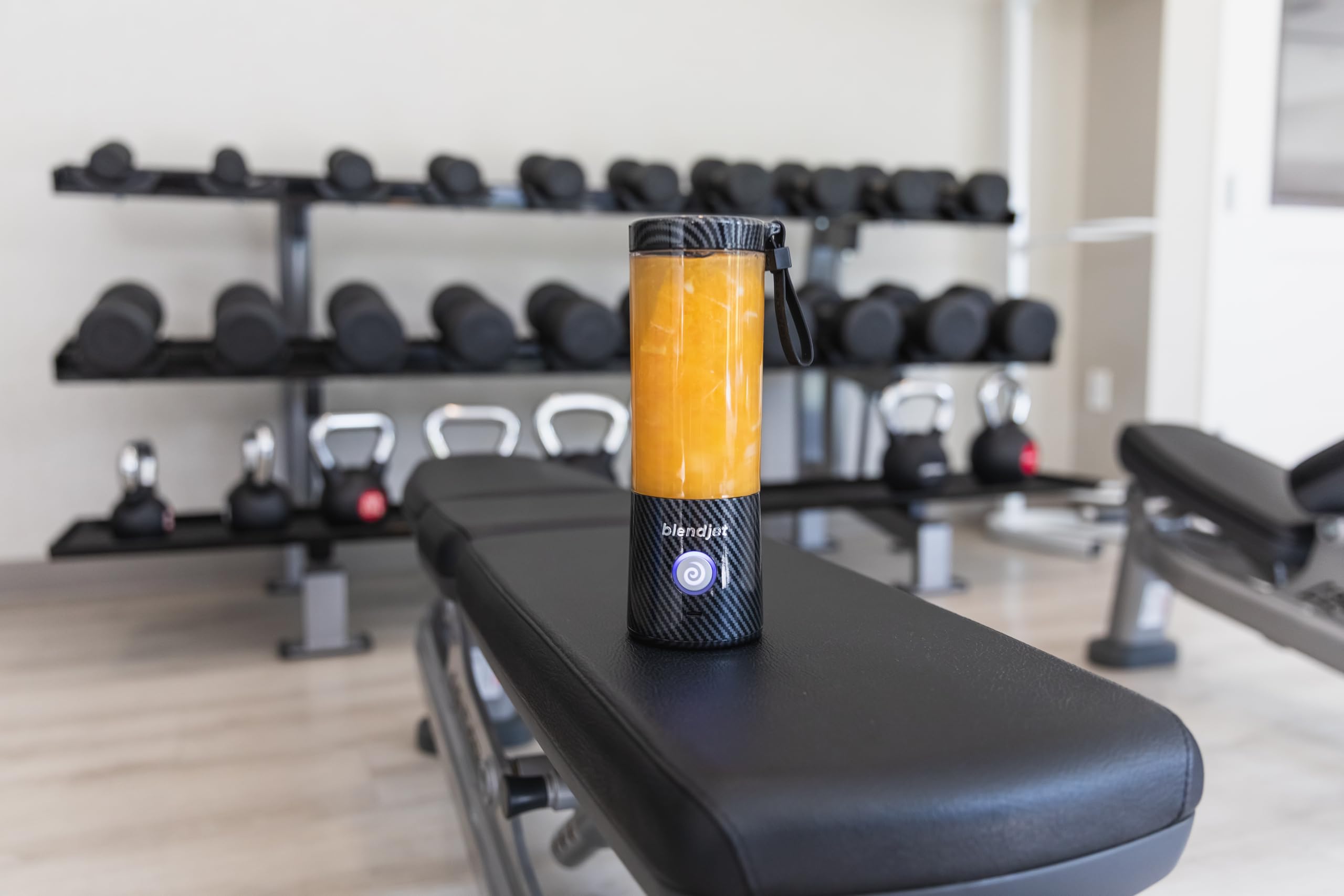 BlendJet 2 portable blender in a sleek carbon fiber design, filled with a fresh smoothie, placed on a gym bench with weights and kettlebells in the background