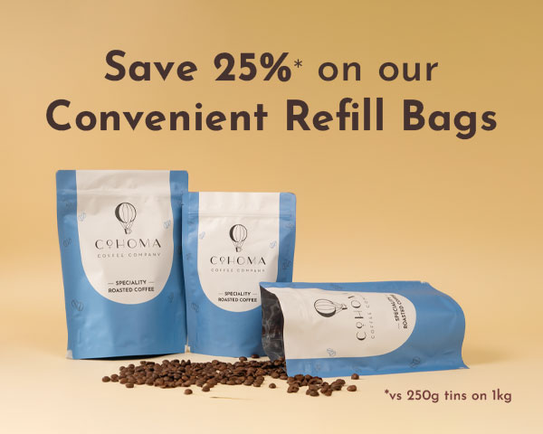 Promotional banner for Cohoma Coffee showcasing blue refill bags of specialty roasted coffee with scattered coffee beans on a beige background